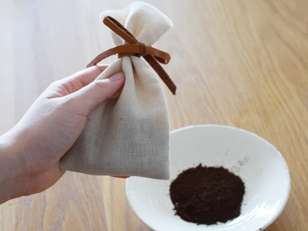 túi thơm vải canvas chứa bã cà phê khô đã buộc dây da, bên cạnh là đĩa đựng bã cà phê, minh họa cách tự làm túi khử mùi bằng bã cà phê để hút ẩm và loại bỏ mùi hôi trong tủ lạnh, giày hoặc xe hơi